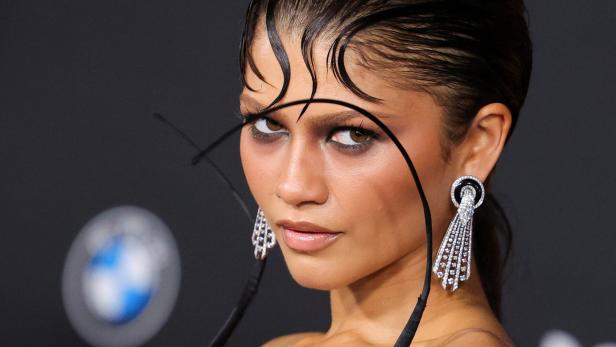 Zendaya blickt mit nassem Haar-Styling und auffälligen Ohrringen vor einem dunklen Hintergrund mit BMW-Logo in die Kamera.