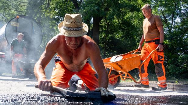 Drei Arbeiter asphaltierten eine Straße, einer kniet vorn mit Kelle, ein anderer schiebt eine Schubkarre, im Hintergrund steht ein Fahrzeug.