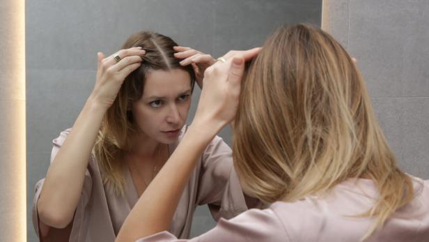 Portrait of a beautiful young woman examining her scalp and hair in front of the mirror, hair roots, color, grey hair, hair loss or dry scalp problem