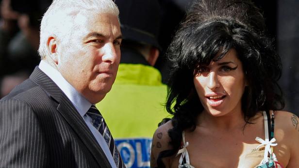 Amy Winehouse steht neben ihrem Vater Mitch im Anzug, während im Hintergrund ein Polizist in neongelber Weste zu sehen ist.
