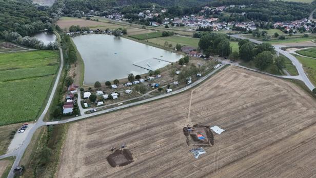 Luftaufnahme eines Dorfes mit See, Feldern, Hügeln und einer archäologischen Ausgrabung im Vordergrund.