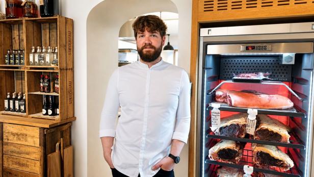 Ein bärtiger Mann in einem weißen Hemd steht vor einem Dry-Ager-Kühlschrank mit Fleischstücken neben einem Regal mit Stieglbräu-Flaschen.