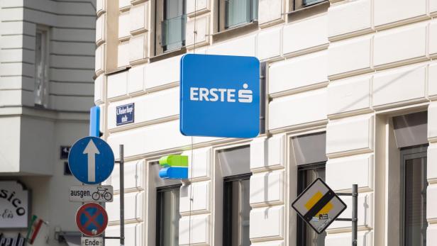 Straßenecke mit ERSTE-Bankenschild und mehreren Verkehrsschildern an einer hellen Hausfassade.