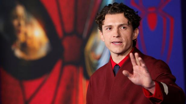 Tom Holland trägt ein weinrotes Sakko vor einem Spider-Man-Hintergrund und hebt grüßend die Hand.