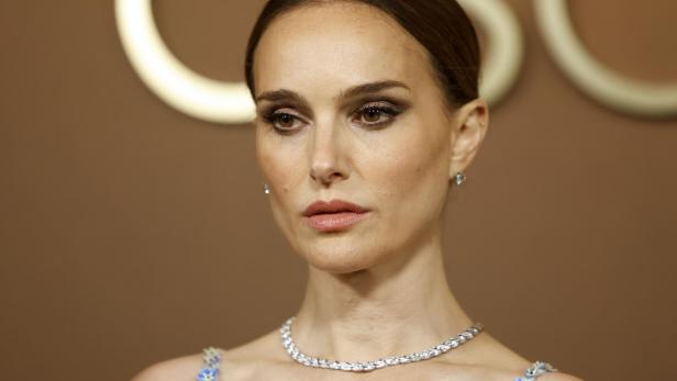 Natalie Portman trägt ein elegantes Collier und ein verziertes Kleid vor einem goldfarbenen Hintergrund.