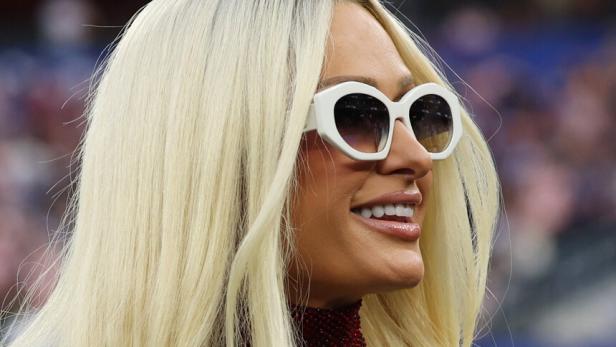 Paris Hilton trägt langes blondes Haar, eine weiße Sonnenbrille und ein glitzerndes Oberteil.