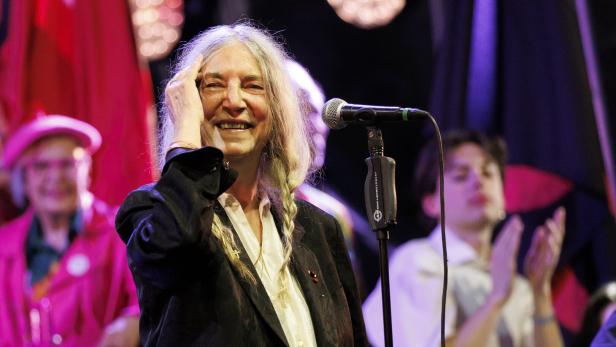 Patti Smith lächelt auf einer Bühne vor einem Mikrofon, während sie sich mit der Hand durch ihr langes graues Haar fährt.