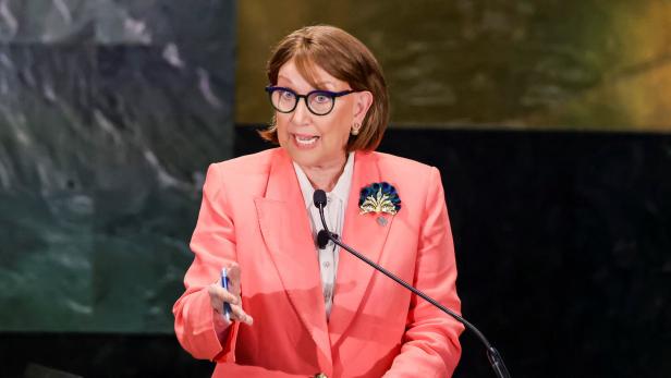 Ältere Frau in rosa Blazer spricht am Rednerpult und gestikuliert mit einem Stift.