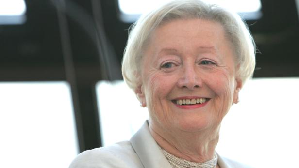 Lächelnde Karin Kathrein mit weißem Haar in heller Jacke und Perlenkette vor hellem Hintergrund.