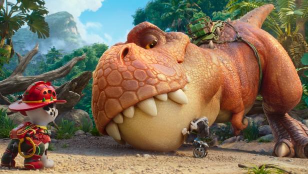 Marshall von der Paw Patrol steht in einem Dschungel vor einem großen T-Rex, an dessen Kopf ein kleiner Hund mit Rollstuhl lehnt.