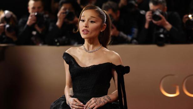 Ariana Grande trägt ein schwarzes schulterfreies Kleid und eine Halskette vor einer Kulisse aus Fotografen.