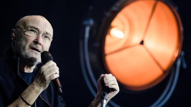 Phil Collins singt vor einem großen, orange leuchtenden Scheinwerfer in ein Mikrofon.