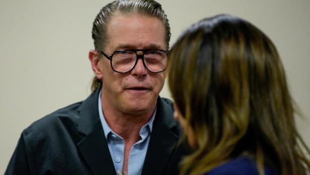Stephen Baldwin mit zurückgegeltem Haar und schwarzer Brille spricht mit einer Frau, von der nur der Hinterkopf und die Schulter zu sehen sind.