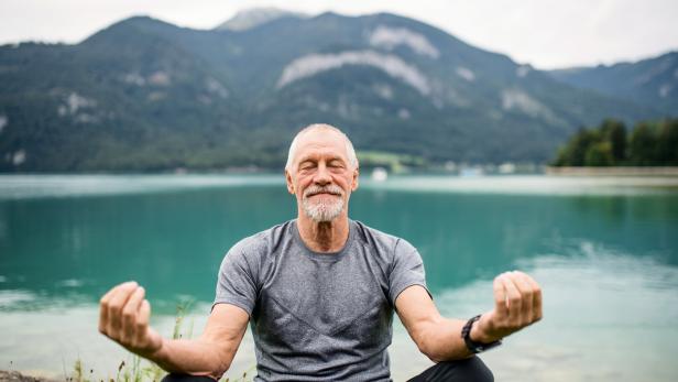 A senior man pensioner sitting by lake in nature, doing yoga exercise.