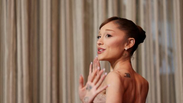 Ariana Grande blickt über die Schulter und hebt die Hand vor einem hellen Vorhang.