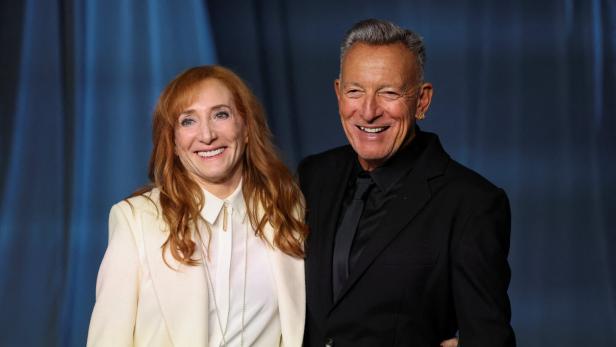 Patti Scialfa und Bruce Springsteen lächeln gemeinsam vor einem blauen Hintergrund in die Kamera.
