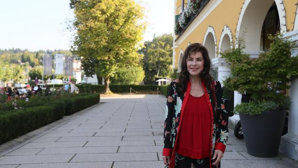 Eine Frau im roten Oberteil und floralen Mantel steht vor einem gelben Gebäude mit Bögen.