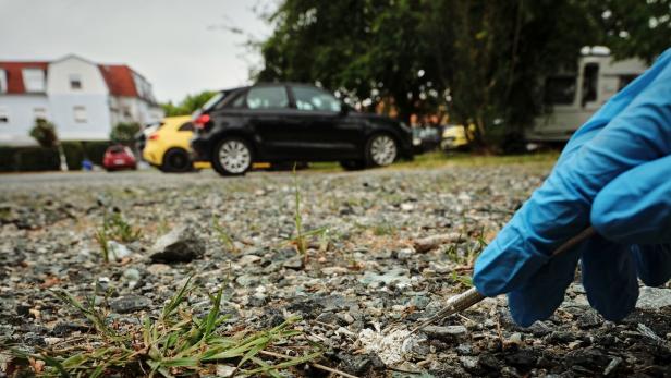 Handschuhhand spritzt weiße Substanz auf den Boden zwischen Gras und geparkten Autos.
