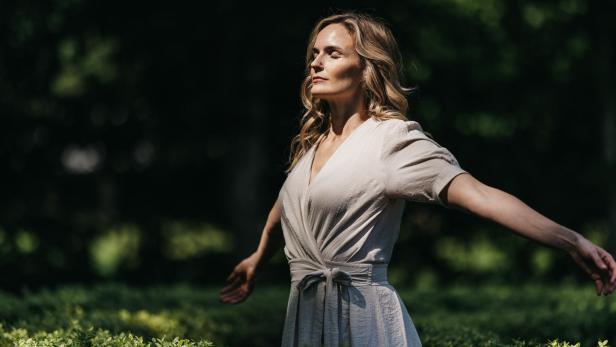 Eine Frau mit blondem Haar steht in einem hellen Kleid mit geschlossenen Augen und ausgebreiteten Armen in einem sonnigen Garten.
