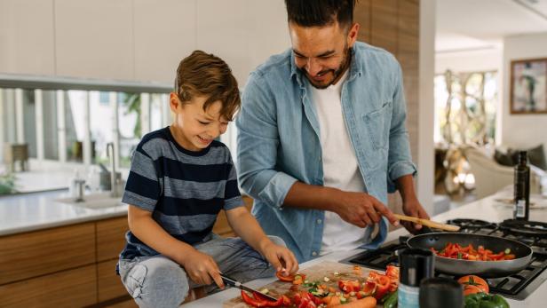 Vater und Sohn kochen gemeinsam in einer modernen Küche, der Junge schneidet Gemüse am Brett.