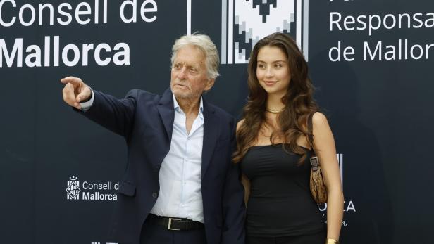 Michael Douglas steht vor einem Banner des Consell de Mallorca und zeigt mit der Hand in die Ferne, während er die Hand Tochter Carys Zeta Jones hält.