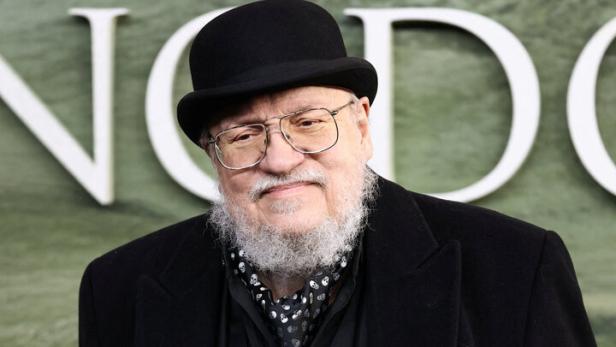 George R. R. Martin trägt einen schwarzen Mantel, eine Weste, eine Melone und eine Brille vor einem grünen Hintergrund.