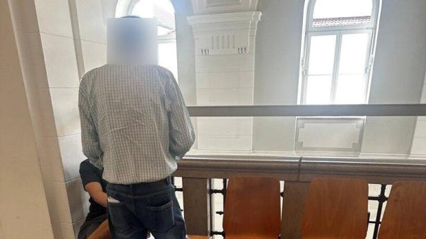 Zwei Männer stehen in einem hellen Flur neben einer Sitzreihe mit Holzstühlen.