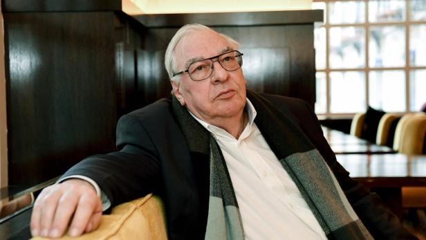 Älterer Mann mit Brille und Schal sitzt in einem Restaurant und blickt nachdenklich zur Seite.