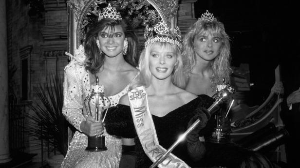 Die Miss World 1987, Ulla Weigerstorfer, sitzt mit zwei weiteren Finalistinnen in Abendkleidern und Kronen auf einer Bühne.
