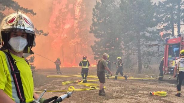 Feuerwehrkräfte bekämpfen einen Waldbrand mit Schläuchen und Ausrüstung, während im Hintergrund Flammen und Rauch aufsteigen.