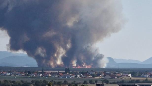 Großbrand mit dichtem schwarzem Rauch über Feldern nahe einer Ortschaft und Bergen im Hintergrund.