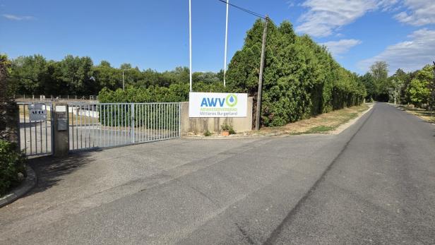 Einfahrt zu einer Anlage mit Schild „AWV Abwasserverband Mittleres Burgenland“, Gittertor, Straße und Bäumen unter blauem Himmel.