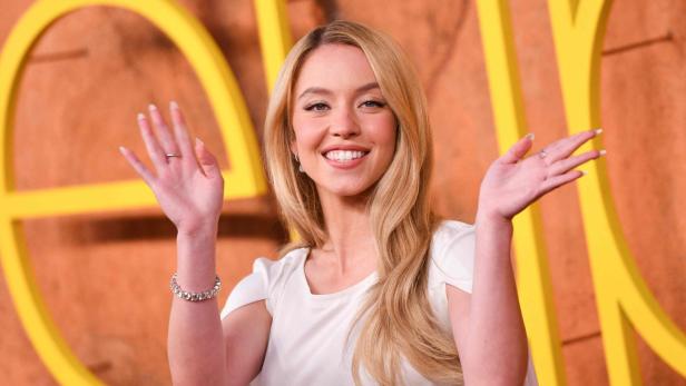 Sydney Sweeney lächelt in die Kamera und winkt mit beiden Händen vor einem gelben Hintergrund.