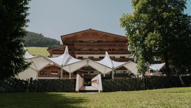 Hotel mit Holzfassade und Sonnensegeln vor grüner Bergkulisse.