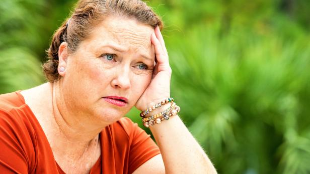 Eine nachdenklich wirkende Frau mit rötlichem Haar und orangefarbenem Oberteil stützt ihren Kopf nachdenklich gegen die Hand.