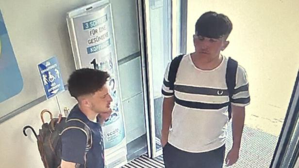 Zwei junge Männer mit Rucksäcken stehen im Eingangsbereich vor einer Glastür.