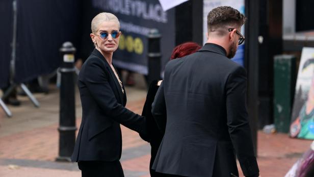 Kelly Osbourne in einem schwarzen Anzug und mit blauer Sonnenbrille geht neben einer Person in einem schwarzen Anzug durch Brindleyplace.