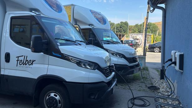 Zwei weiße Elektro-Lieferwagen der Bäckerei Felber stehen an einer blauen Wand und werden an Ladestationen aufgeladen.