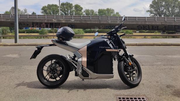 Ein schwarzes Honda-Motorrad mit einem auf dem Sitz liegenden Helm steht an einer Straße vor einer mit Graffiti verzierten Brücke.