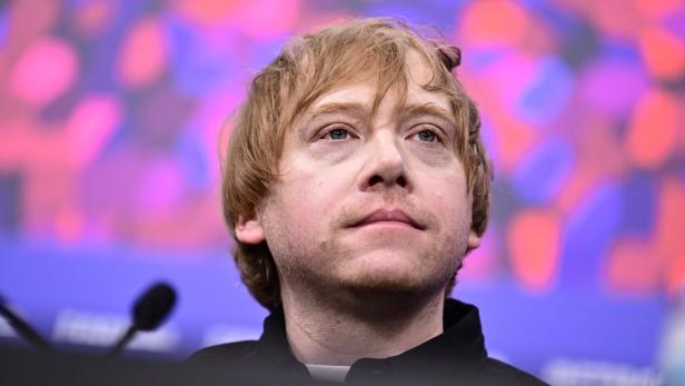 Rupert Grint blickt nachdenklich vor einem violetten Hintergrund mit unscharfen Mustern.