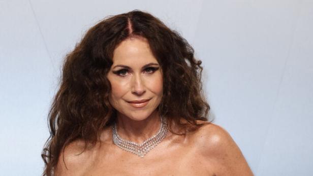 Minnie Driver trägt ein trägerloses rotes Kleid und eine auffällige Diamantkette vor einem hellen Hintergrund.