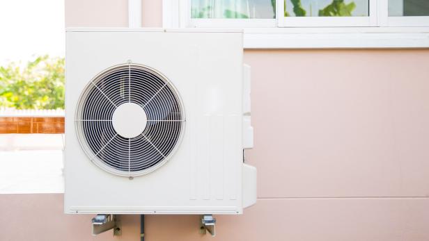 Ein weißes Außengerät einer Klimaanlage ist an einer hellrosa Hauswand unter einem Fenster montiert.