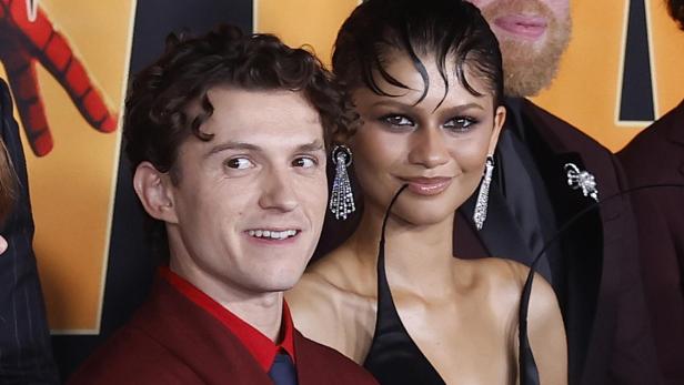 Tom Holland in einem weinroten Anzug und Zendaya in einem schwarzen Kleid posieren gemeinsam vor einem Hintergrund.