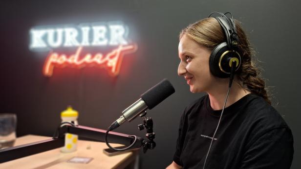 Eine Frau mit Kopfhörern spricht vor einem Mikrofon in einem Studio mit einem leuchtenden „KURIER podcast“-Schriftzug im Hintergrund.