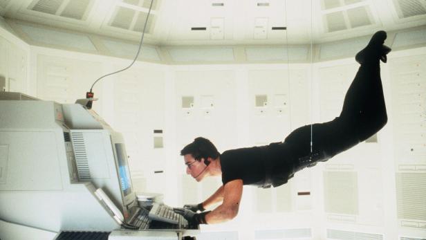 Tom Cruise schwebt in Mission: Impossible an Seilen befestigt in einem hellen Raum und tippt konzentriert auf einer Computertastatur.