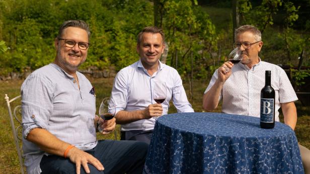 Drei Männer sitzen draußen an einem Tisch, trinken Rotwein und lächeln in die Kamera.