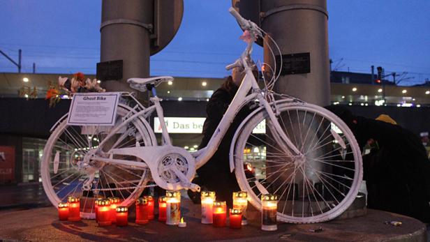 Weiße Gedenkfahrrad-Skulptur mit Kerzen am Fuß einer Brücke bei Dämmerung.