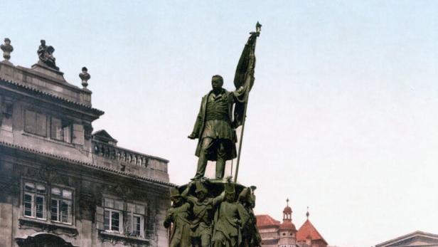 Denkmal eines Generals auf hohem Sockel in einem historischen Prager Platz, umgeben von alten Gebäuden und Passanten.