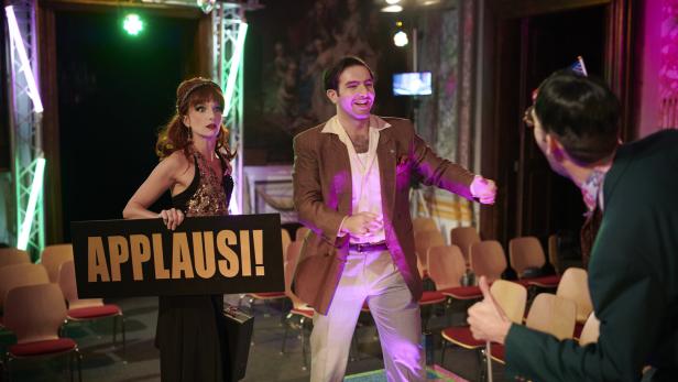 Personen auf einer Bühne bei einer Show, eine Frau hält ein „APPLAUS!“-Schild, ein Mann spricht mit Publikum.