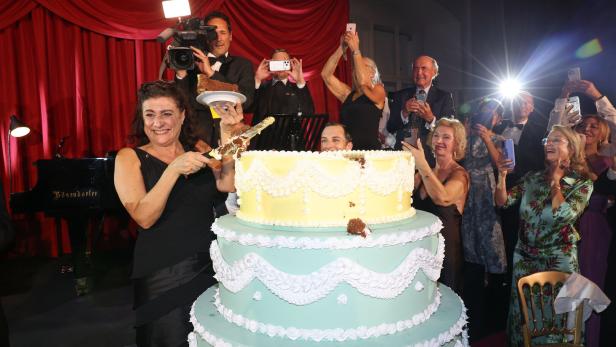 Eine Frau schneidet vor rotem Vorhang eine riesige Torte an, umgeben von applaudierenden Gästen und Fotografen.